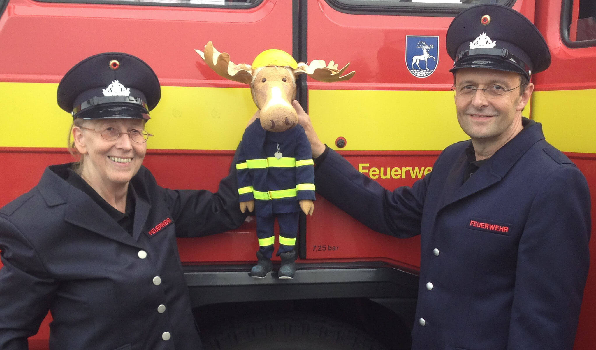 Foto: Doro Wellfonder und Wolfgang Kaup-Wellfonder stehen mit Feuerwehr-Uniformen vor einem Feuerwehrauto und halten den Feuerwehrelch Finn fest.